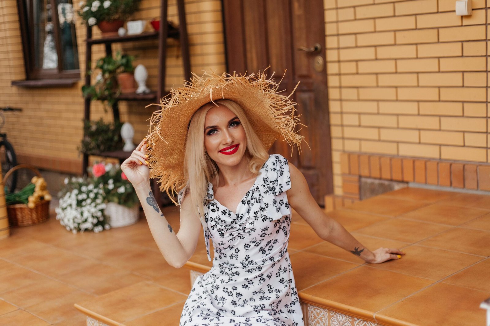 A cheerful woman wearing a straw hat and white sundress, sitting on steps outdoors with floral background.